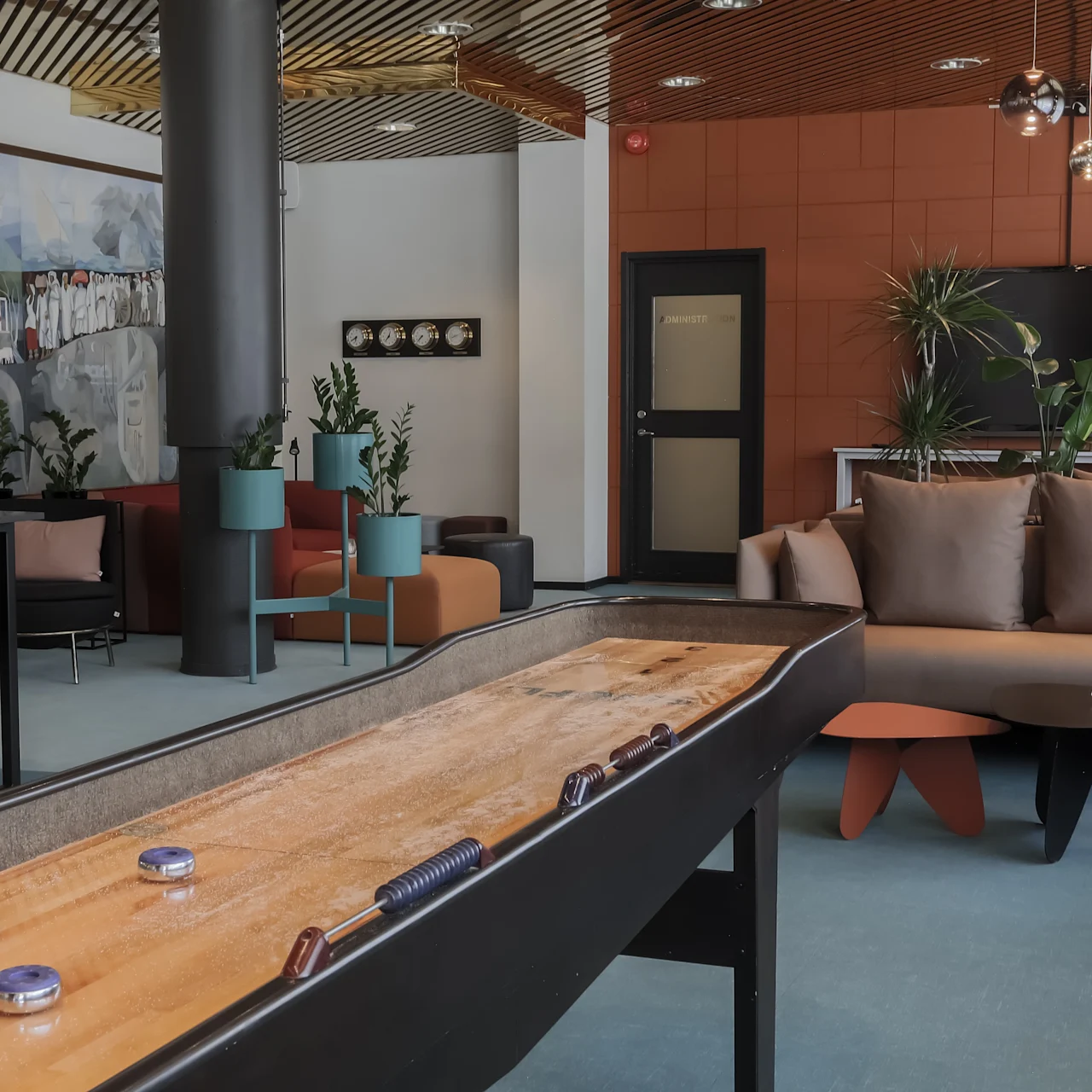 Shuffleboard table with pucks sits idle in a spacious, modern lounge, featuring cozy seating, indoor plants, abstract art on the wall, a TV, and pendant lights.