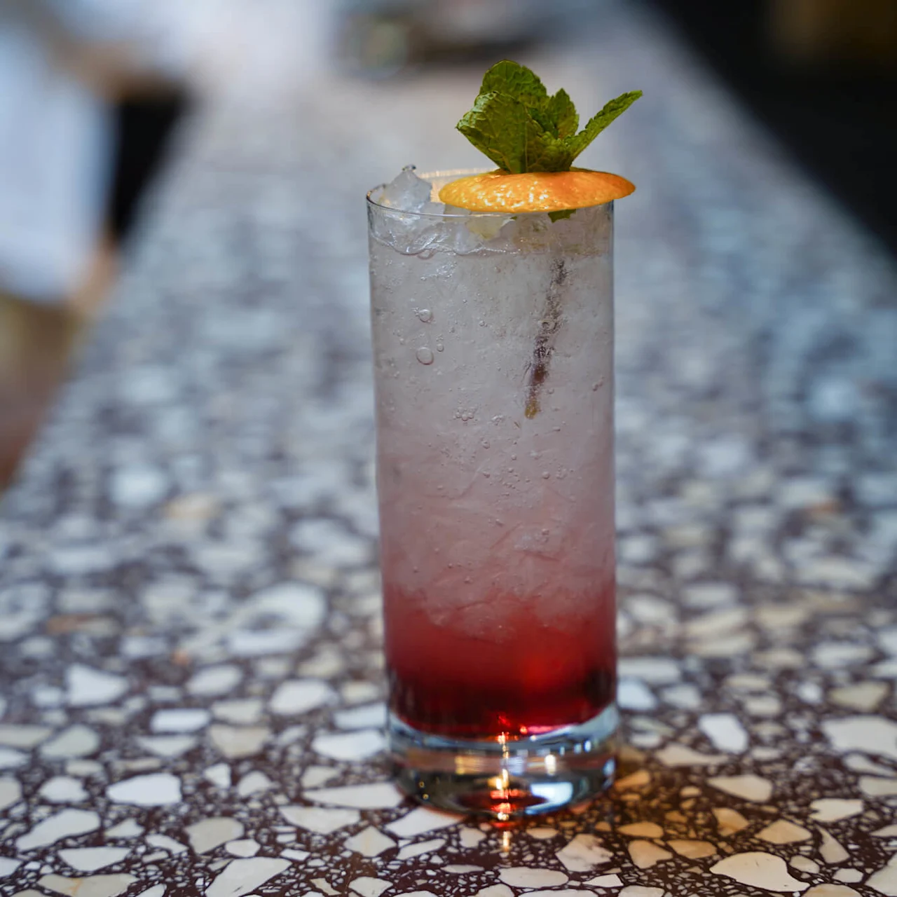 A colorful gradient cocktail on a mosaic table, garnished with mint and an orange slice.