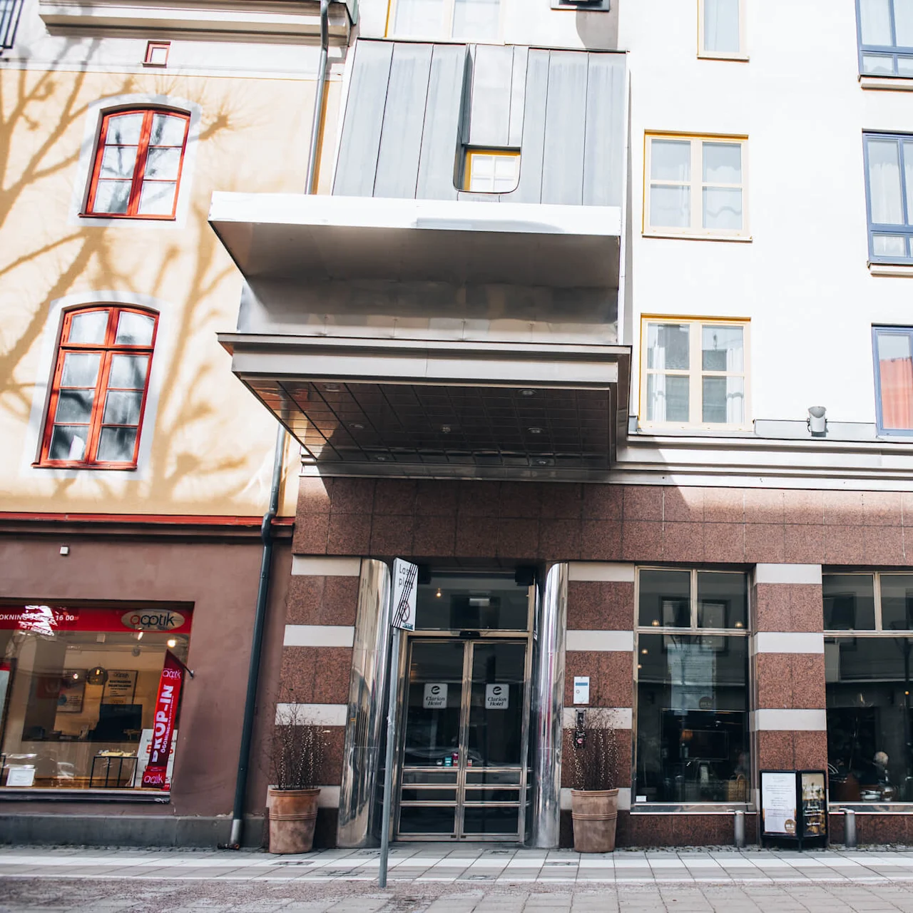 A multi-story building facade with modern glass doors serves as an entrance. Surrounding it, shops and large windows; signage "Lastplats" on a streetlamp in an urban street setting.