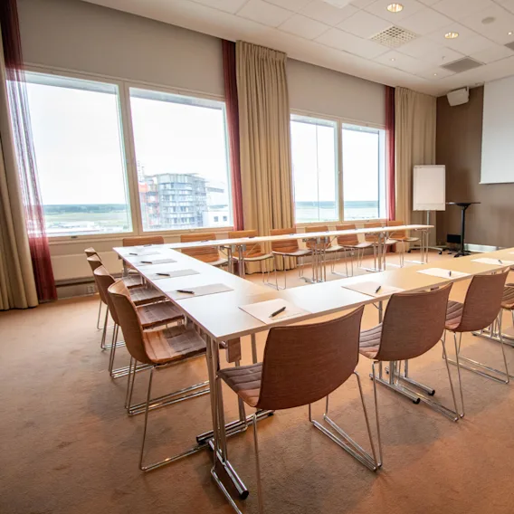 Bord och stolar arrangerade i en U-form, vända mot en stor skärm och ett blädderblocksställ i ett ljust konferensrum med stora fönster och beige gardiner.