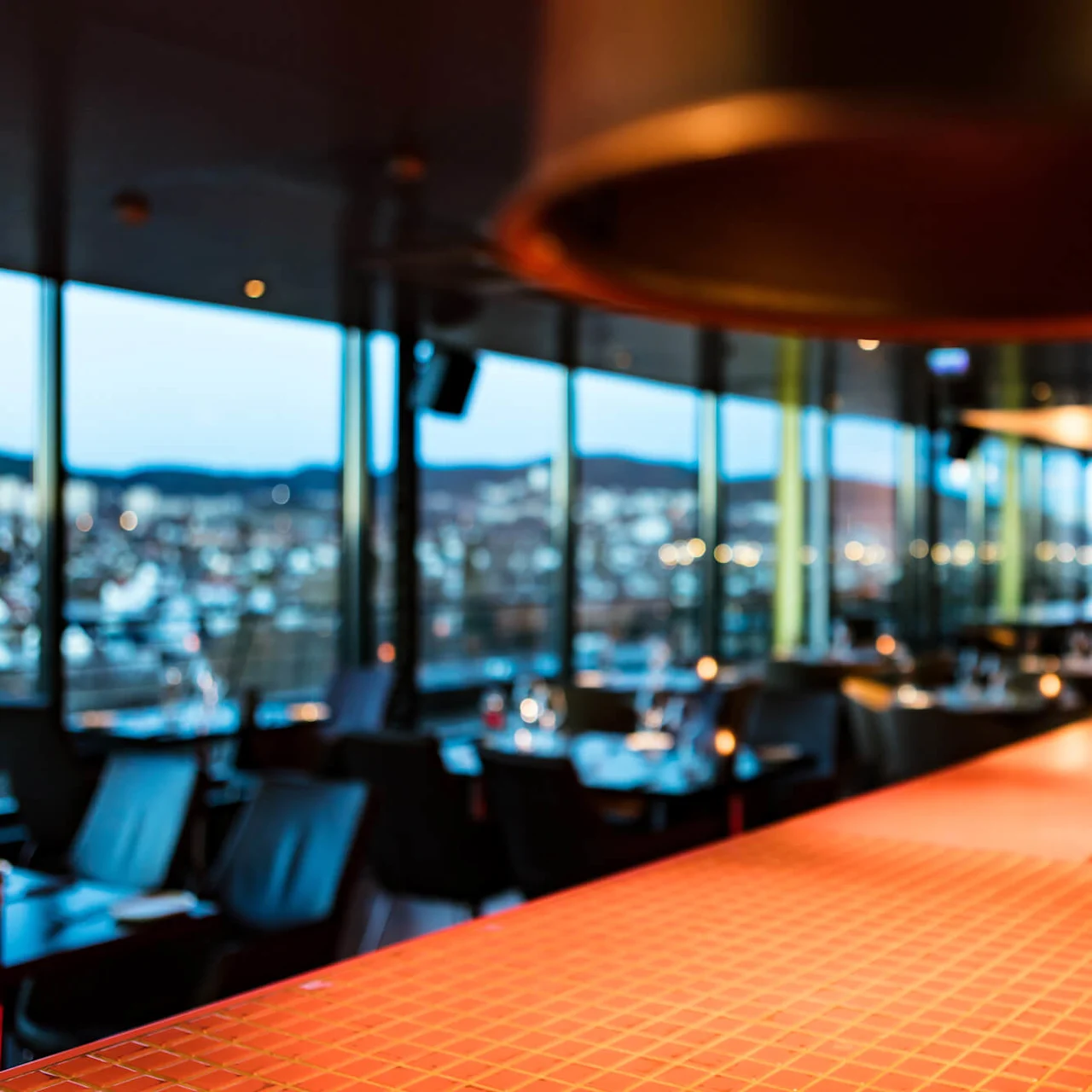 A copper-colored hanging lamp is suspended over a tiled counter in a modern, dimly-lit restaurant with large windows offering a blurred city skyline view in the background.
