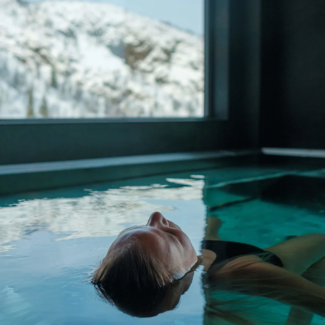 A person floats calmly on their back in an indoor pool, with snowy mountains visible through a large window.