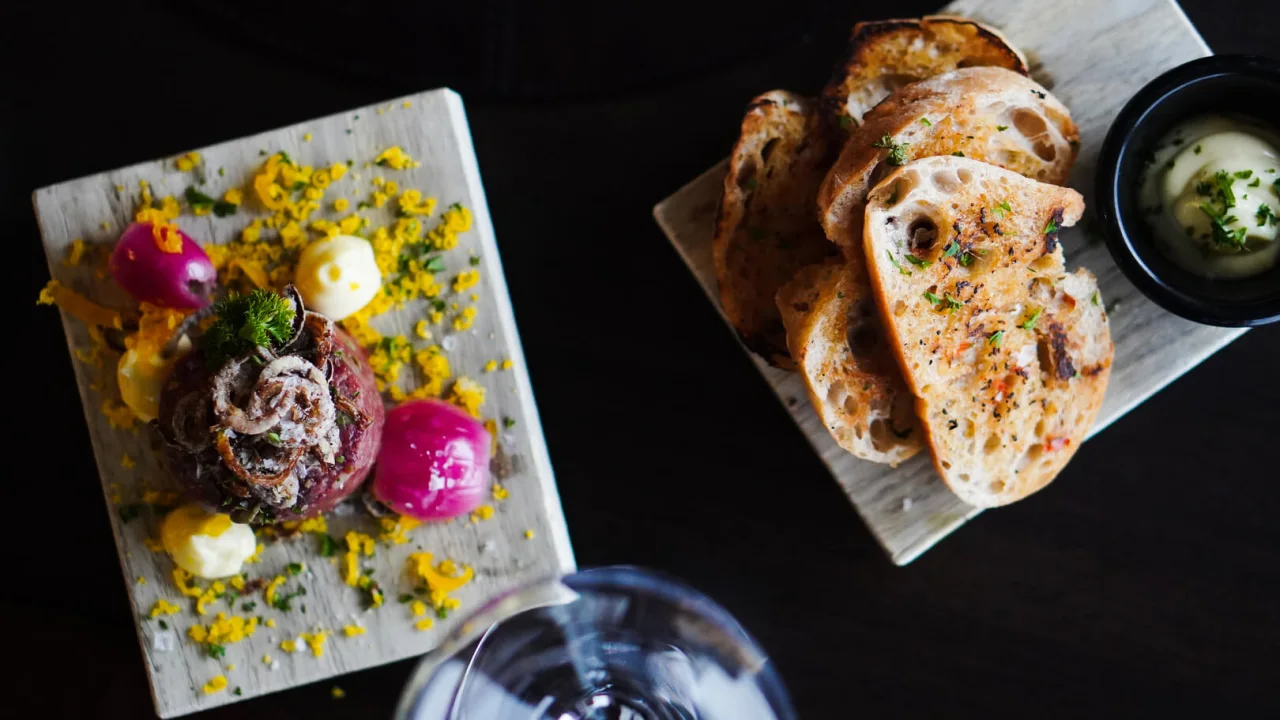 Gourmet dish served on a wooden board with herb-crusted beef, colorful accompaniments, and sliced bread on a dark table.