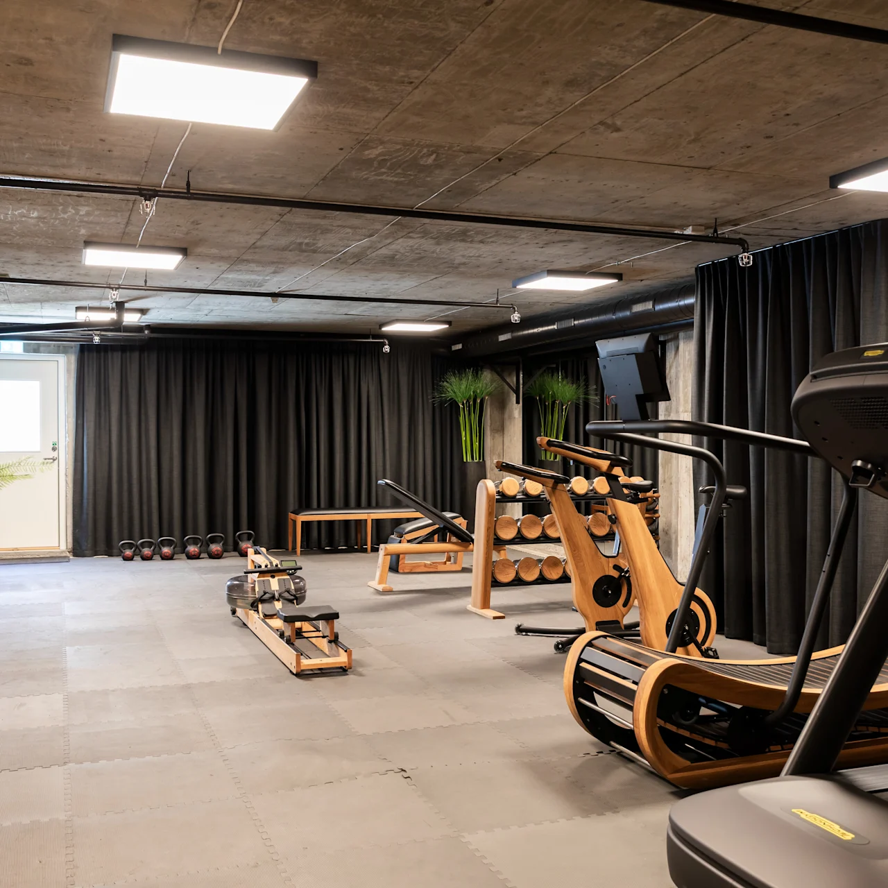 Løbebånd står stille i et moderne træningsstudie med træ-træningsudstyr, mørke gardiner og potteplanter, hvilket skaber et elegant, minimalistisk træningsrum.