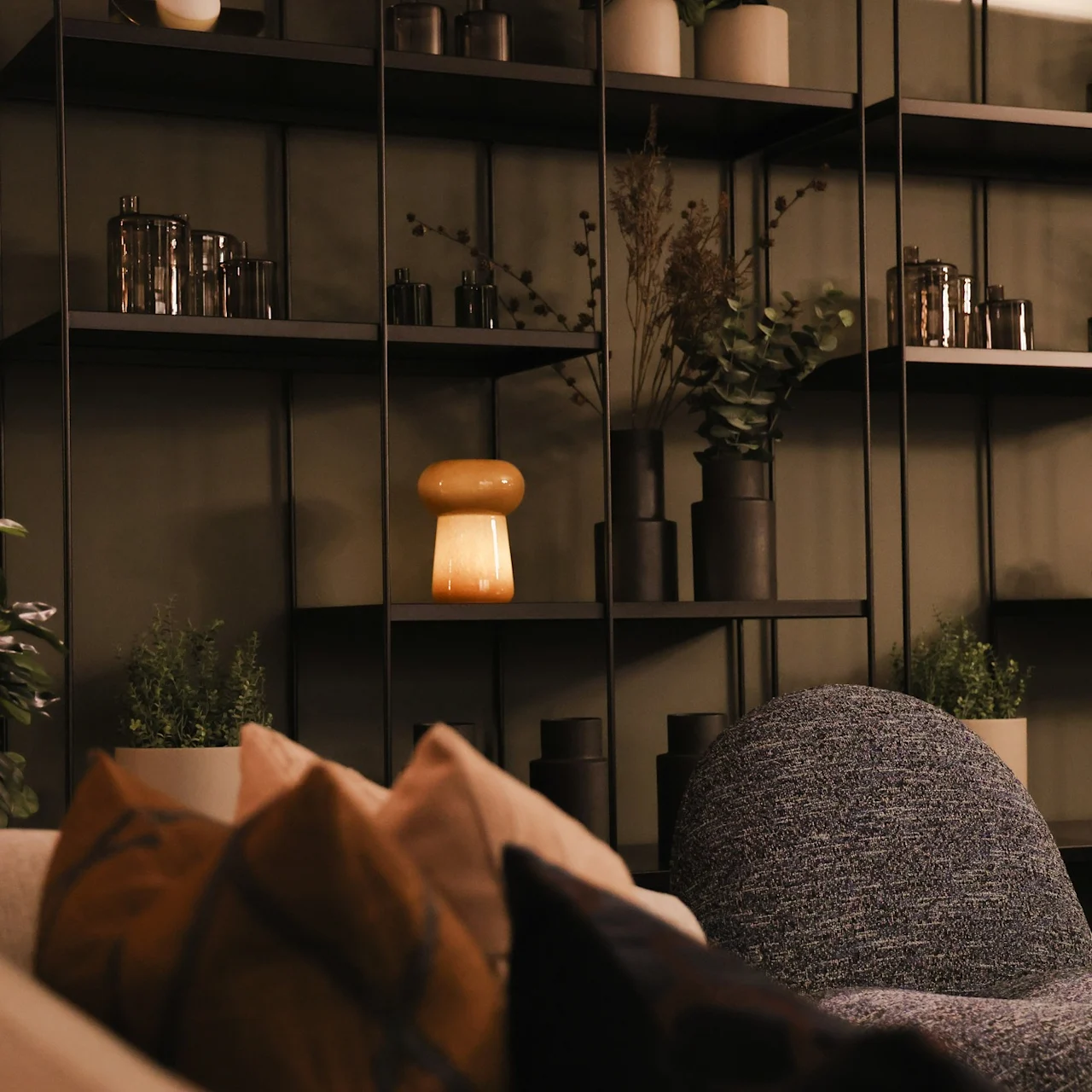 A glowing mushroom-shaped lamp sits on a dark shelf, surrounded by decorative vases and plants in a cozy, modern living room with soft chairs and pillows.