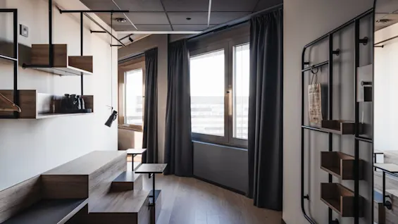Room with modern wooden furniture, built-in shelving, and a desk lamp. Dark curtains partially cover large windows, allowing light into the space. The walls are light-colored, enhancing the minimalist design.
