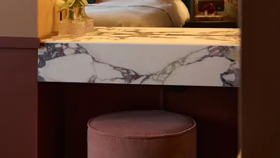A pink velvet stool with fringe accents stands under a marble vanity with illuminated mirror bulbs. In the background, a bed with white pillows and a decorative headboard is visible.