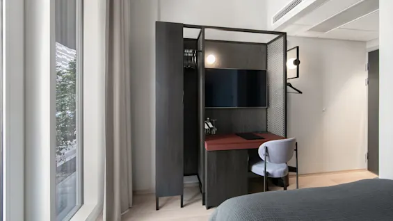 A modern hotel room workspace features a dark wood wardrobe and desk with a mounted TV. A chair is placed at the desk, and large windows with curtains are nearby.