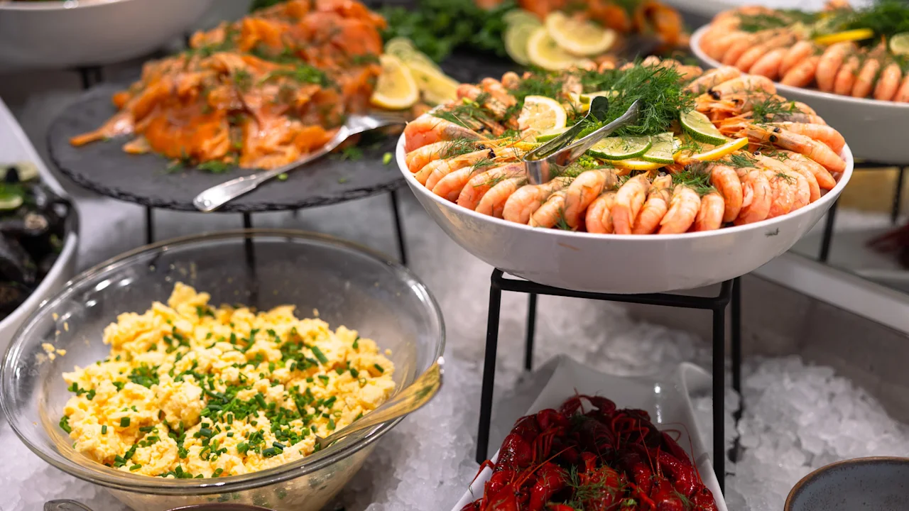 En skaldjursbuffé med räkor garnerade med citron och dill i en vit skål, omgiven av rökt lax, äggröra med gräslök och lysande röda kräftor, på en bädd av is.