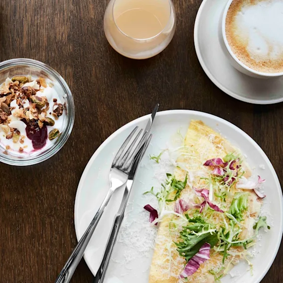 En tallerken med en omelett toppet med grønnsaker og radicchio ligger på et trebord ved siden av en kopp kaffe, et glass juice, yoghurt med granola og et rundstykke.