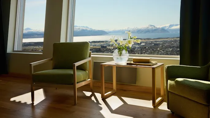 Green armchair sits by a table with a vase of lilies and two books, overlooking a scenic view of mountains and water through large windows.