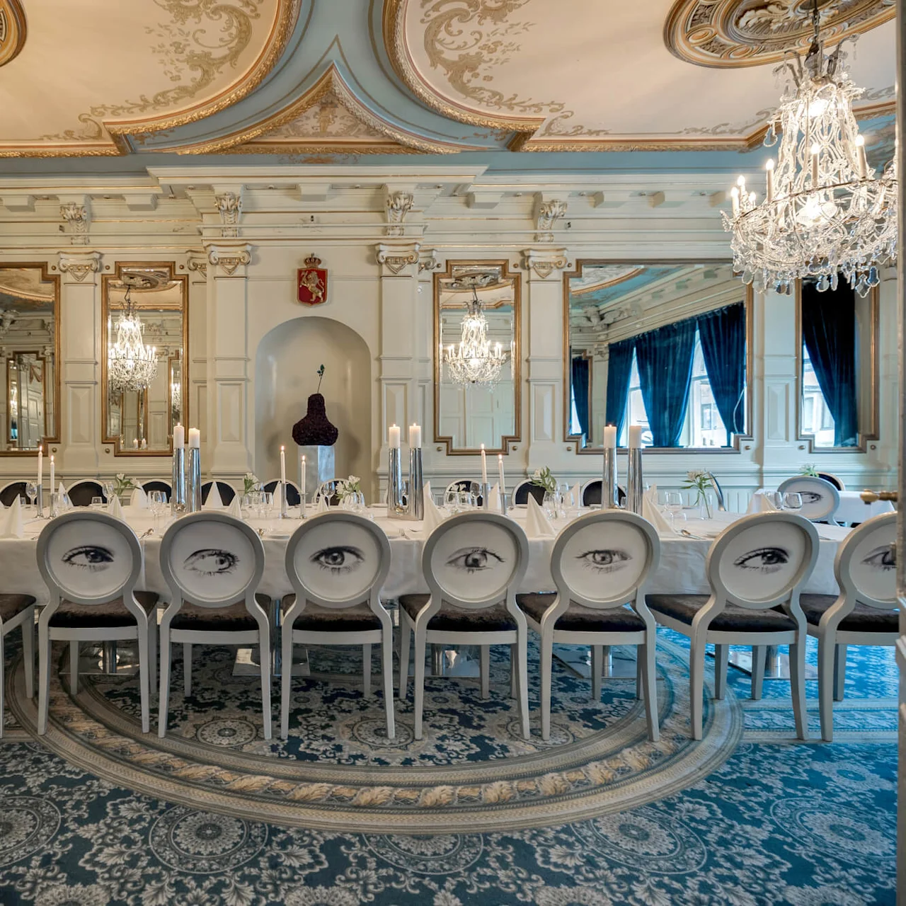 Elegante spisestole med øjendesign på ryggen omgiver et stort bord dækket med hvide duge og stearinlys i et overdådigt rum med lysekroner, udsmykkede spejle og detaljerede lofter.