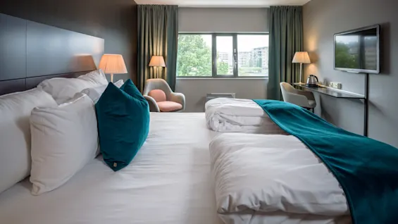 En pænt redt seng med hvidt sengetøj og turkis accenter står i et moderne hotelværelse. Værelset har et vindue med byudsigt, lamper, en stol og et vægmonteret tv.