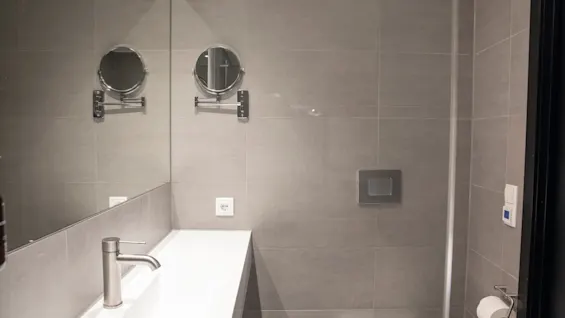 Et rent og moderne bad med grå fliser, en hvit vask, et stort speil og et toalett med en rull toalettpapir.