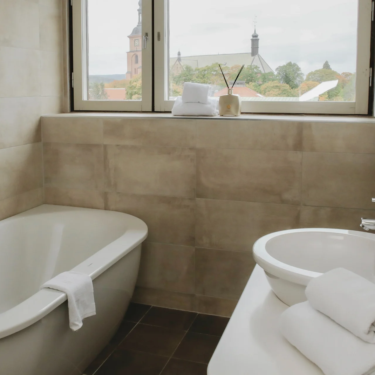 Bathtub with a towel on it and a sink beside, in a tiled bathroom with a window showing church spires and rooftops outside.