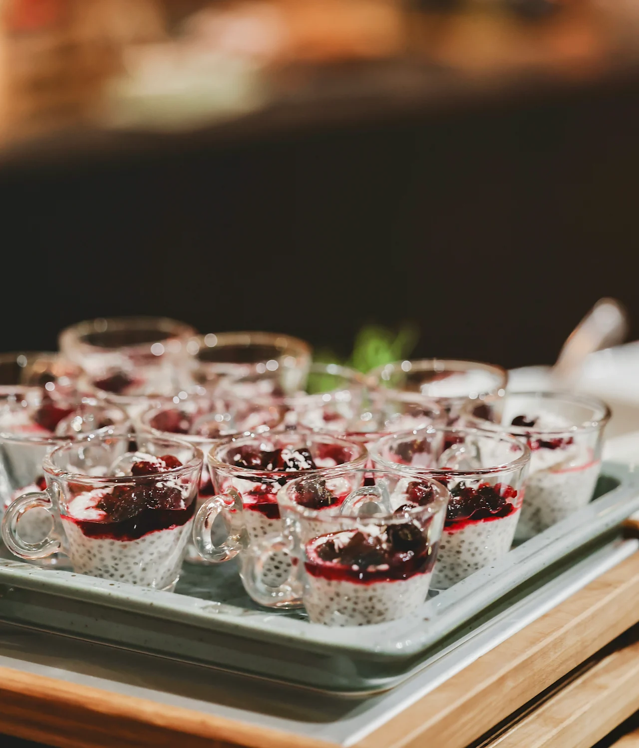 Small glass cups hold chia pudding topped with dark berry sauce, arranged neatly on a tray, all placed on a wooden table in an indoor setting.