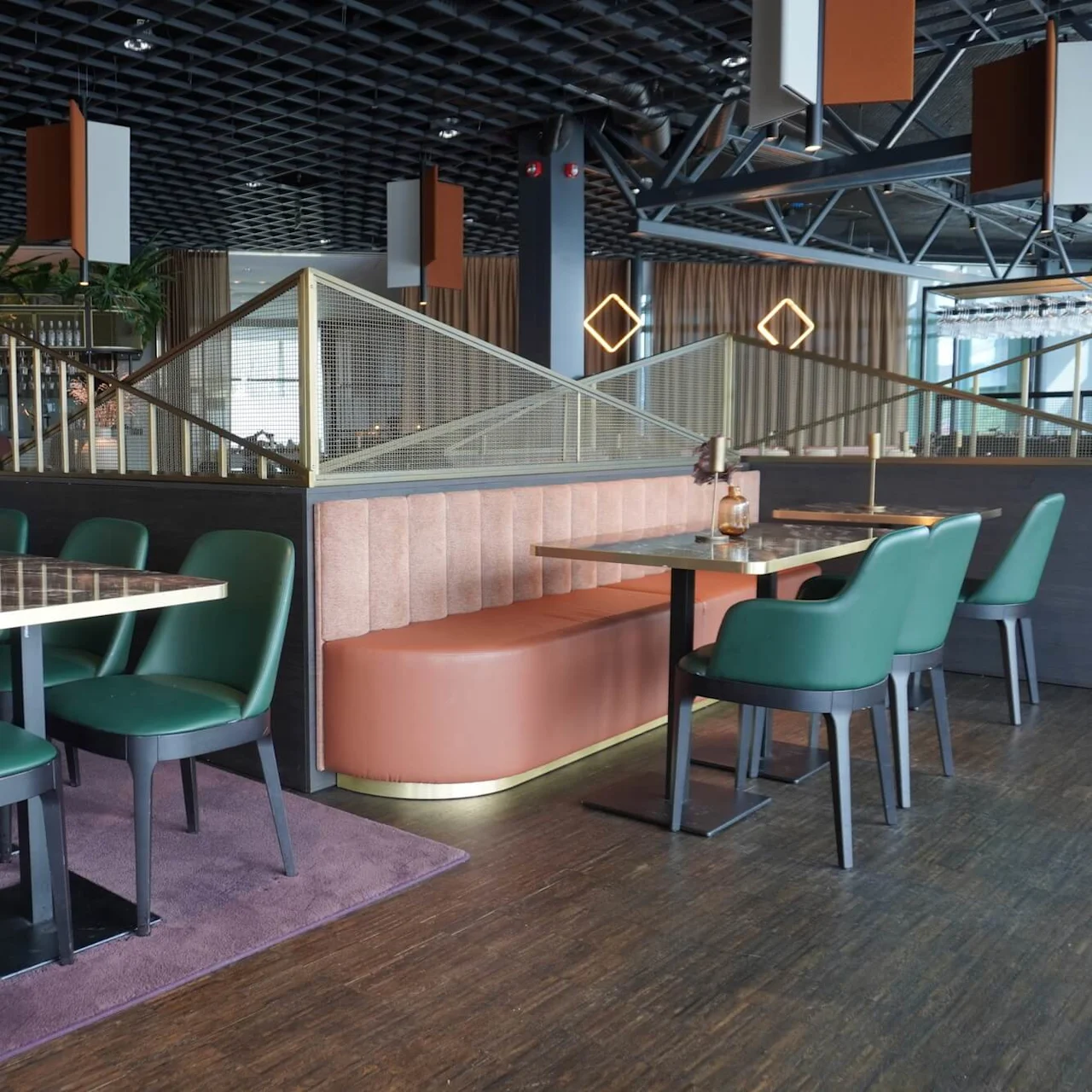 Dining tables with green chairs and brown booths sit in a modern restaurant setting, featuring geometric ceiling decor and warm lighting, complemented by decorative vases and plants.