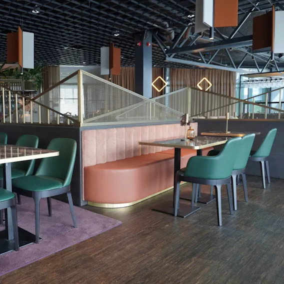 Spisebord med grønne stoler og brune sofaer står i et moderne restaurantmiljø, med geometrisk takdekor og varm belysning, supplert med dekorative vaser og planter.