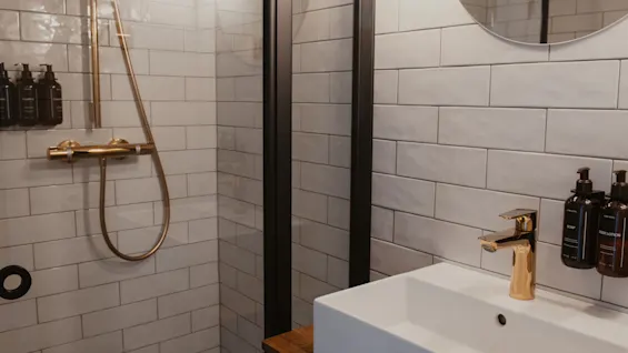 Moderne badeværelse med hvide flisevægge, bruser og vask med guldfarvede armaturer. Flasker mærket SÆBE og BODY LOTION.
