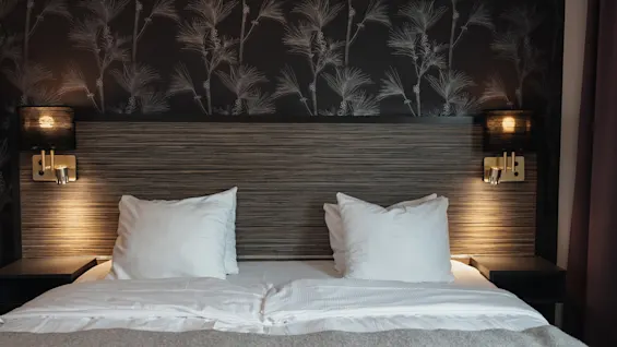 Viihtyisä hotellisänky valkoisilla liinavaatteilla, puinen sängynpääty ja lämmin valaistus yöpöytävalaisimista.