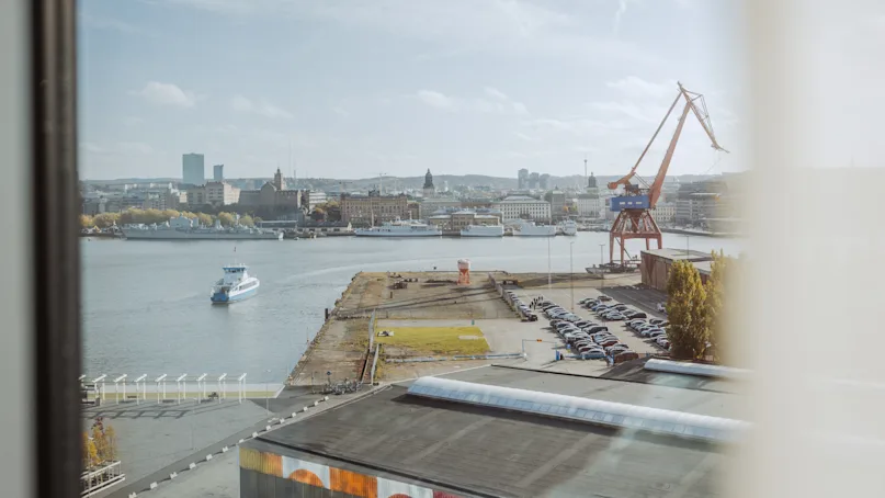 A river with a boat sailing; large crane stands on the dockside; industrial buildings and parked cars visible around; cityscape with various buildings in the background under a partly cloudy sky.