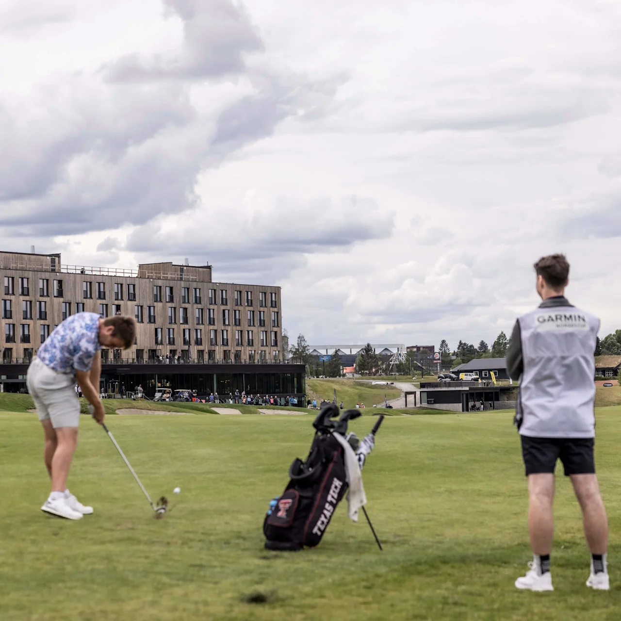 En golfspelare svingar mot en boll på en frodig bana, åskådare observerar; byggnader i bakgrunden.