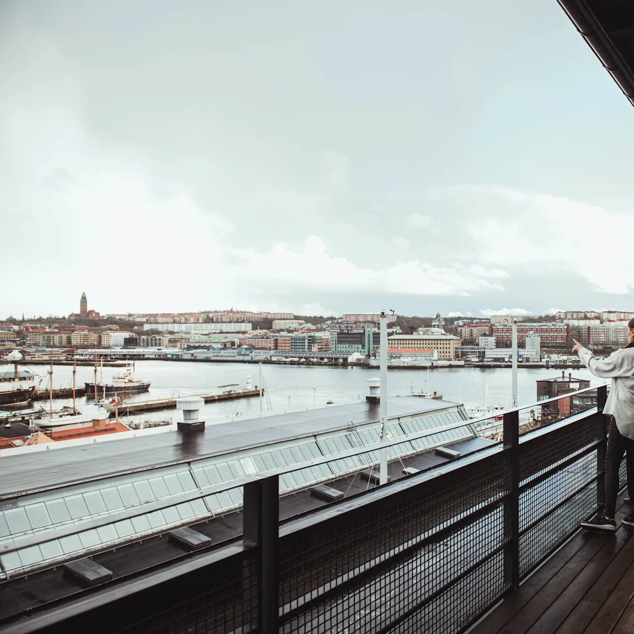 Five people and a dog stand on a balcony, talking and pointing, overlooking a cityscape and harbor with boats and buildings under a cloudy sky.