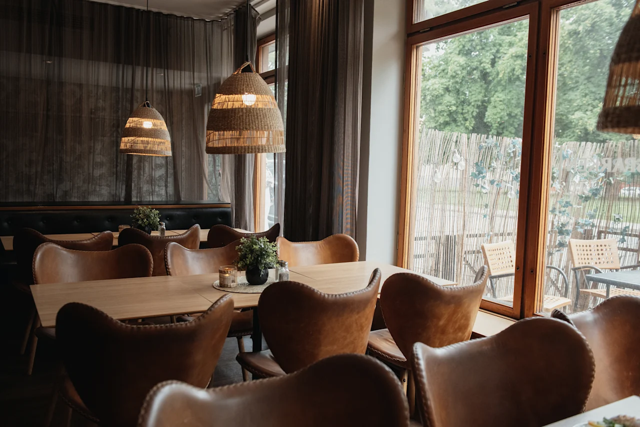 A cozy restaurant interior with warm lighting, wooden tables, and leather chairs, looking out onto an outdoor patio.