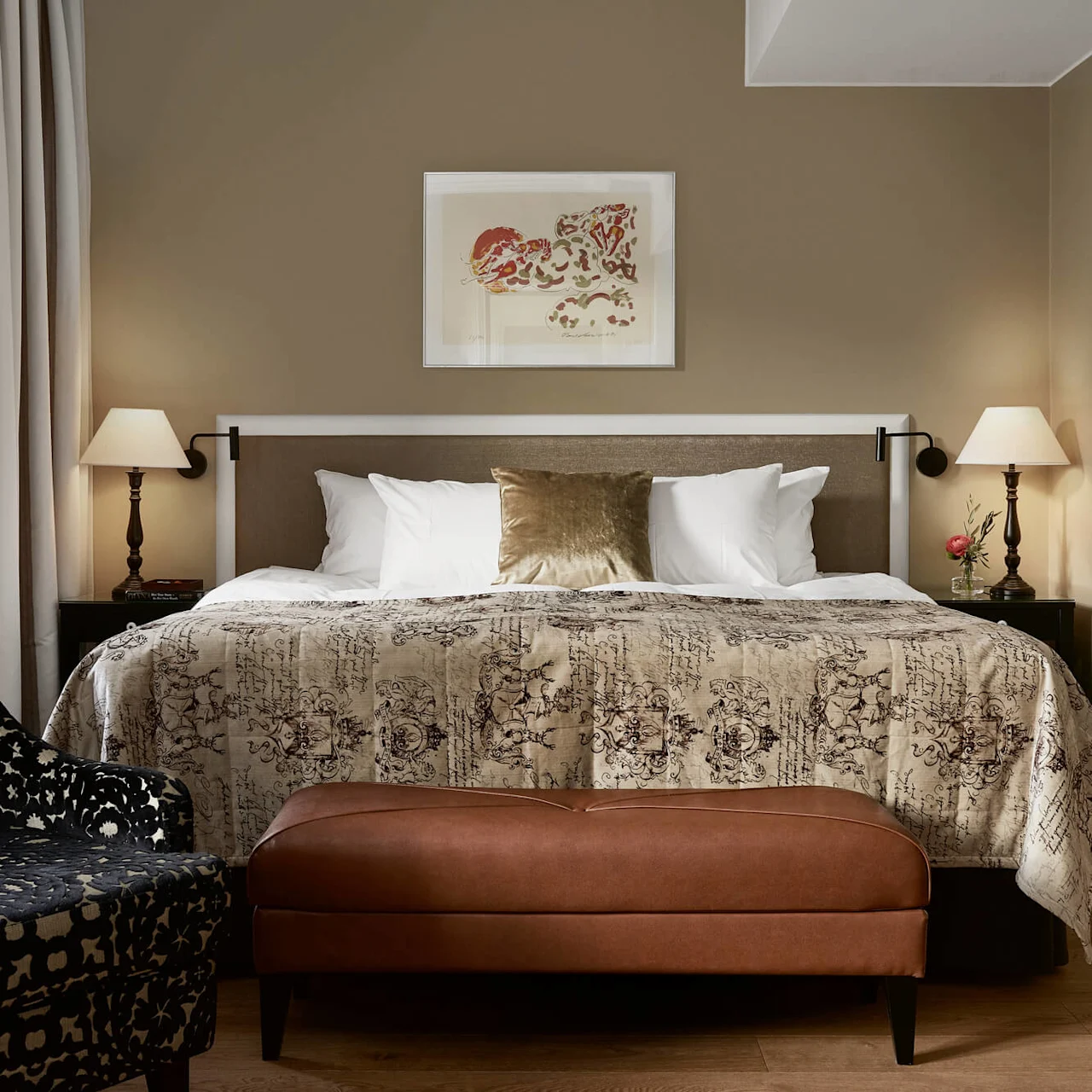 A king-sized bed with patterned bedspread and pillows is flanked by lamps on nightstands. An ottoman sits at the foot of the bed in a softly lit bedroom.