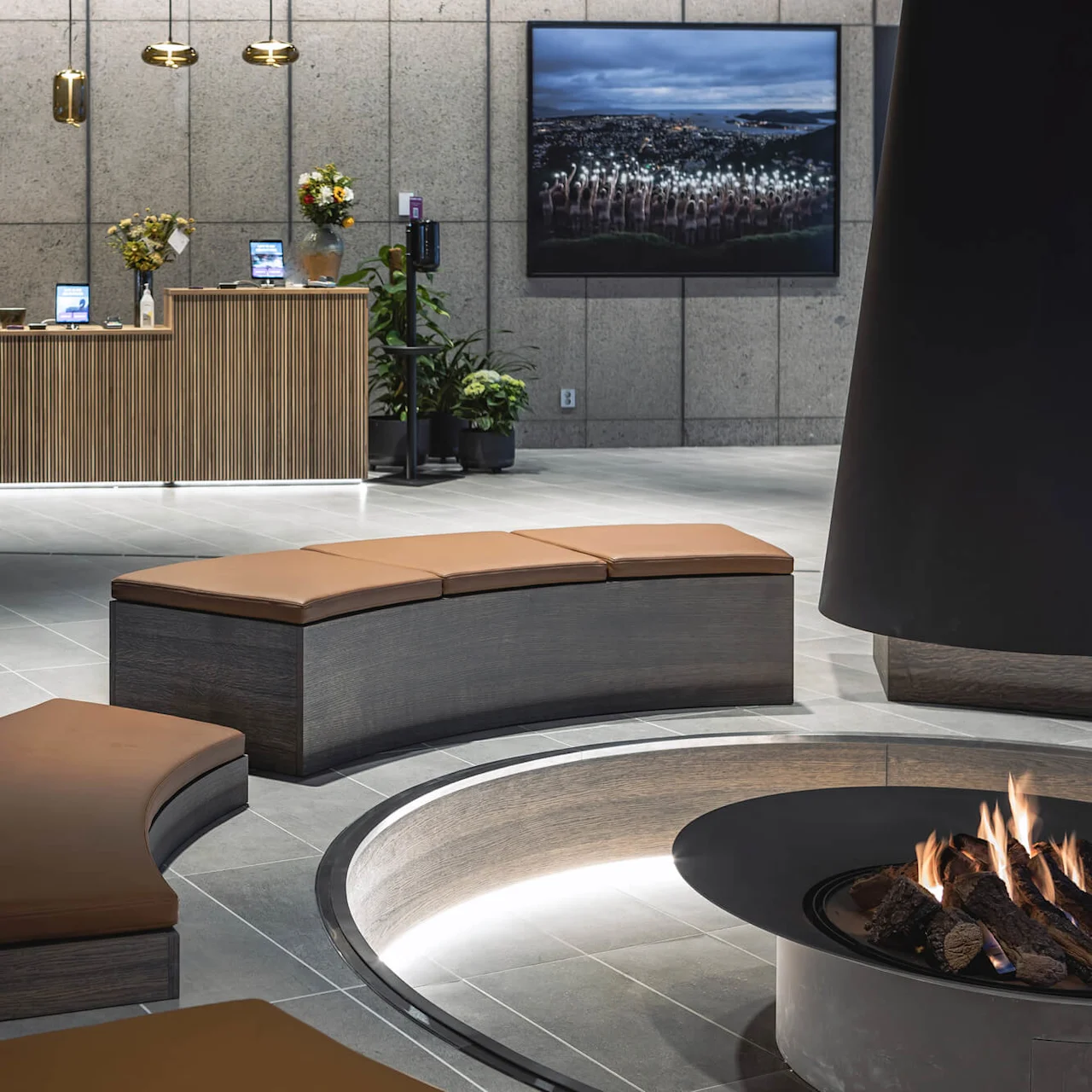 A circular fireplace emits warmth in a modern lobby with curved seating. Wooden reception desks hold flowers and tech devices under pendant lights. A landscape photo decorates the concrete wall.