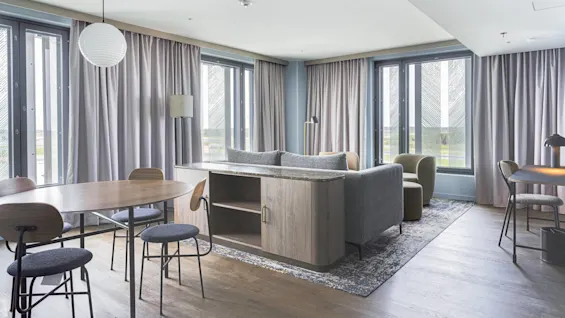 A modern living room features a gray sofa and wooden cabinet, surrounded by light curtains. A round dining table with chairs and a work desk form a cozy, well-lit space.