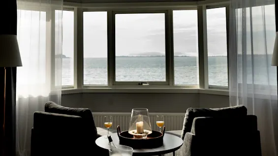 To lænestole vender mod et stort karnapvindue, der vender ud mod havet. Et lille bord har et lys og glas, hvilket skaber en indbydende, afslappet stuestemning.