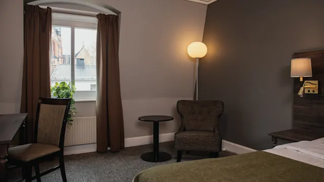 A hotel room features a bed, chair, and round table beside a floor lamp. A window with curtains reveals building rooftops outside.