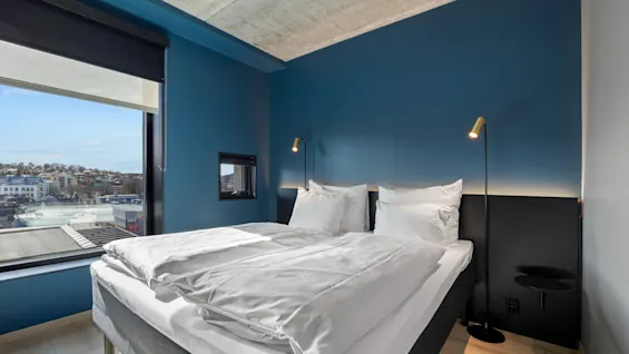 Et moderne hotellrom med en komfortabel seng, blå vegger og et stort vindu med utsikt over byen.
