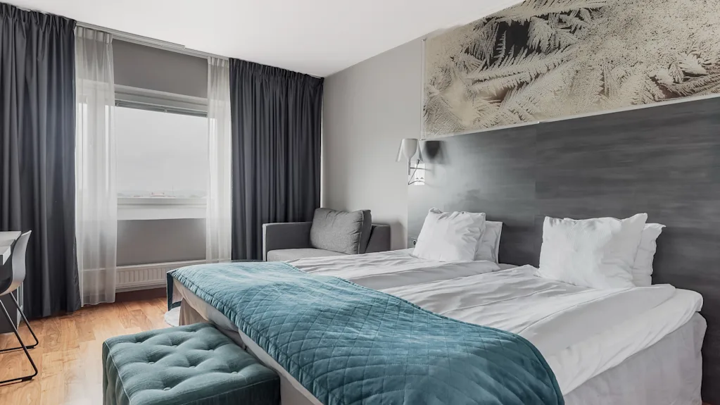 A comfortable hotel room with two beds, a cozy blue throw, and a window offering natural light. Perfect for relaxation.