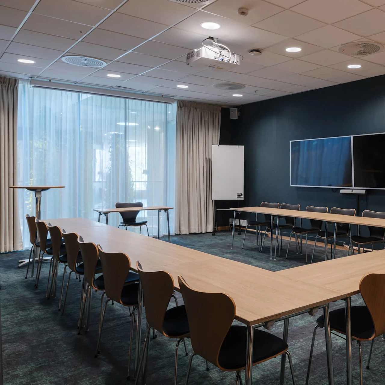 Ett U-format konferensbord med flera stolar står i ett mötesrum. En stor skärm är monterad på väggen, och en whiteboard står bredvid golvlånga gardiner.