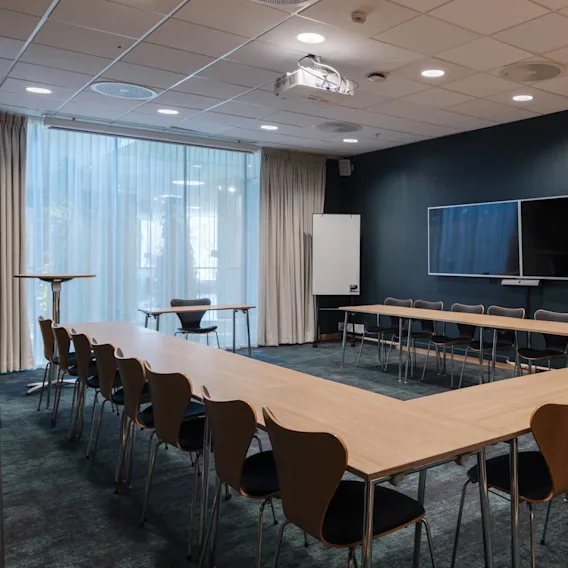 Ett U-format konferensbord med flera stolar står i ett mötesrum. En stor skärm är monterad på väggen, och en whiteboard står bredvid golvlånga gardiner.