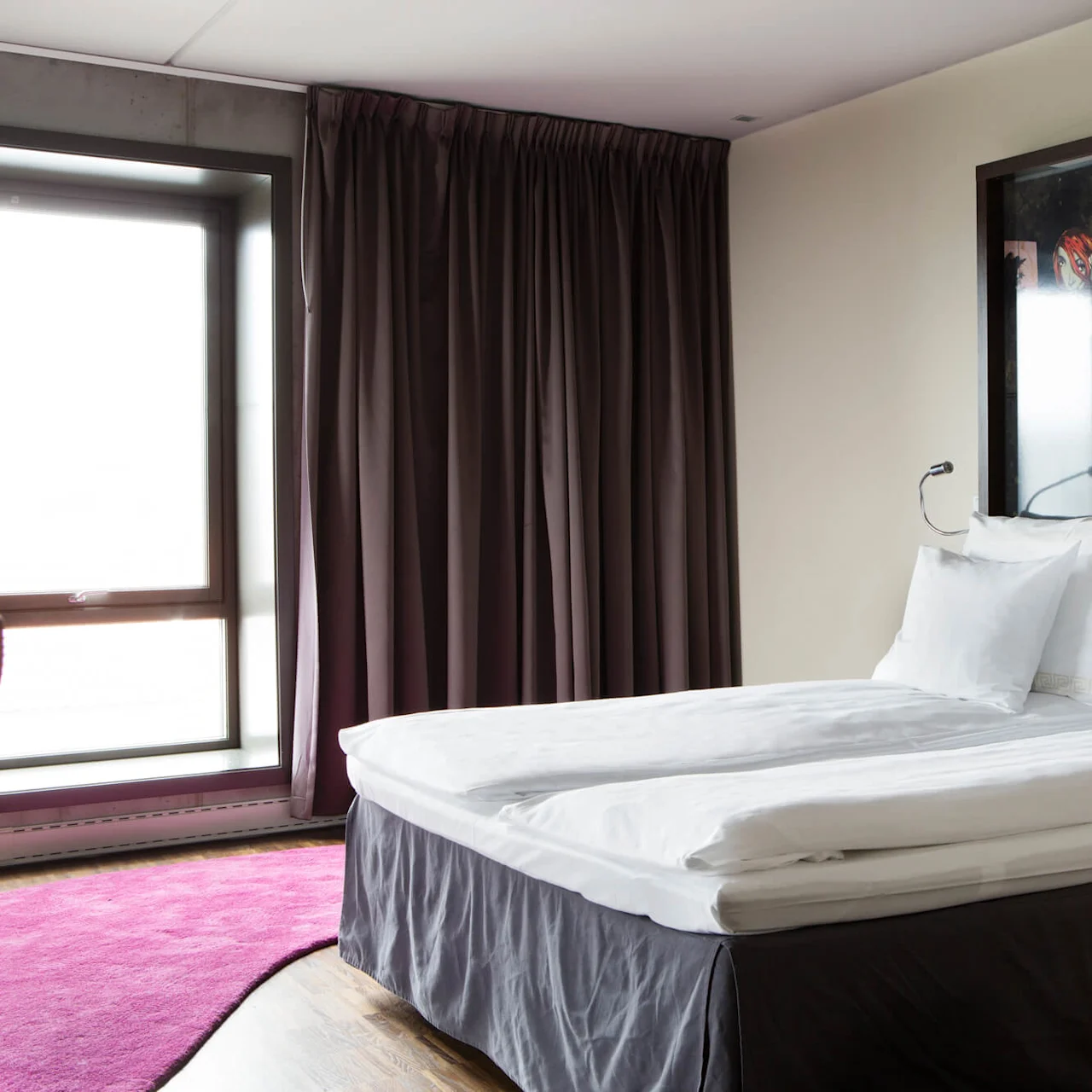 En pent dobbelt seng med hvite laken står i et moderne hotellrom. Bakgrunnen har mørke gardiner og et stort illustrert sengegavl med en rødhåret figur.