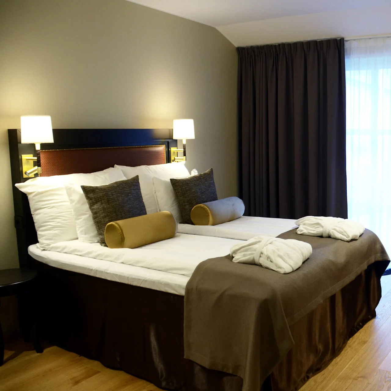 A neatly made bed with pillows, in a well-lit hotel room with curtains, a chair, and bedside lamps.
