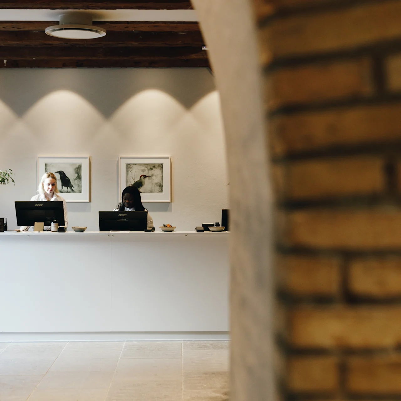 Reception desk with two people working on computers, framed by an archway. The room has white walls, a tiled floor, and two framed artworks on the wall.