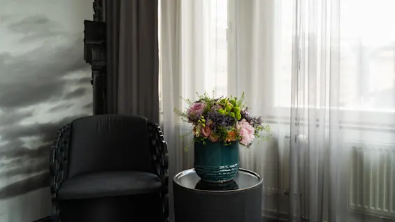 A flower arrangement in a green ceramic vase sits on a round table. Nearby, a dark chair rests against a wall adorned with a cloudy sky and building mural.
