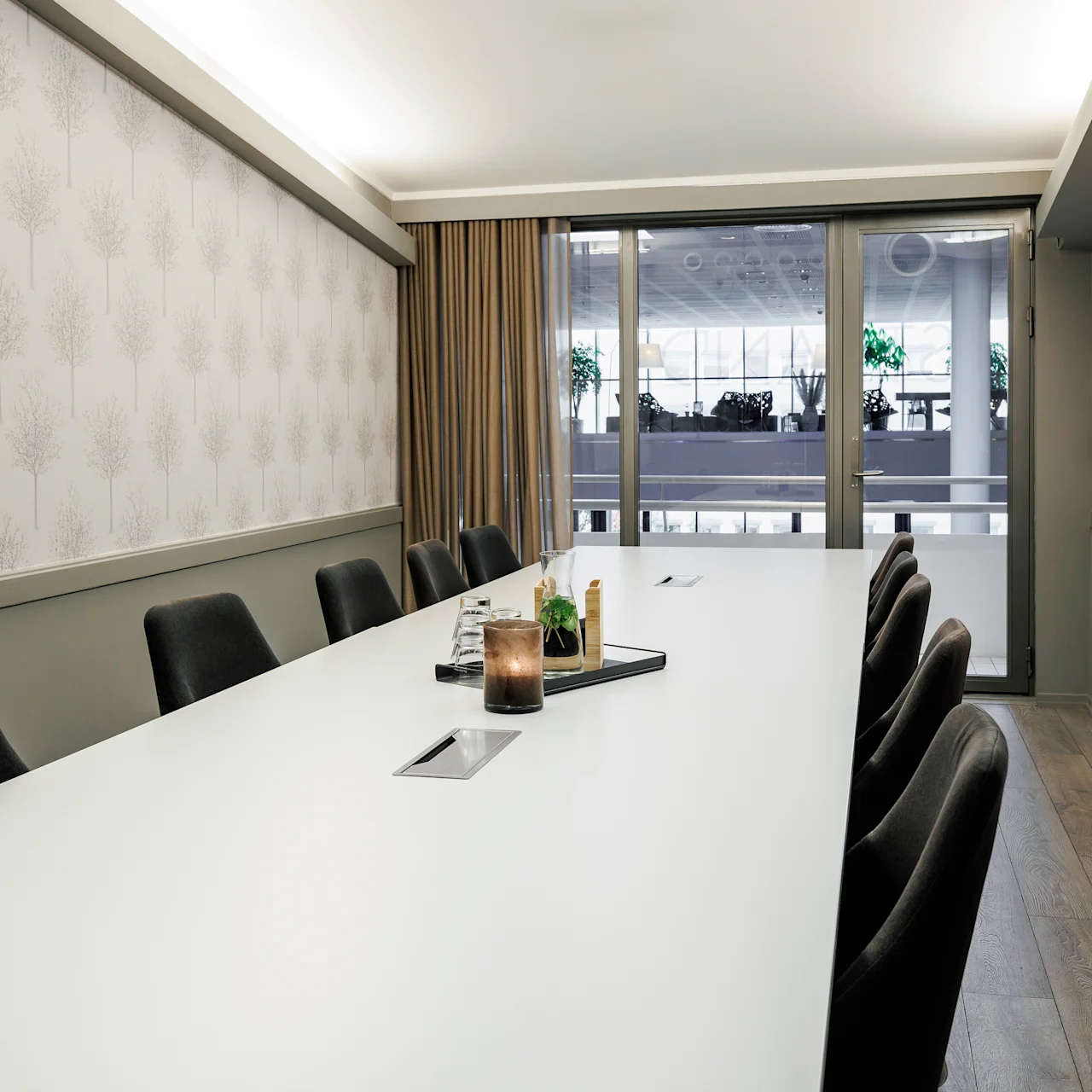 A modern conference room features a long white table surrounded by black chairs. A candle and plant centerpiece adorn the table. Large windows and patterned wallpaper enhance the bright space.