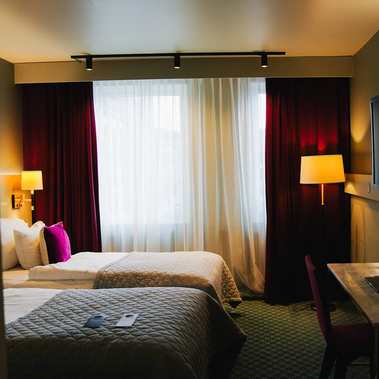 En hotellrum med två sängar med quiltade överkast, ett skrivbord med stol, en väggmonterad TV och ett stort fönster med vinröda gardiner. Belysningen är varm och inbjudande.