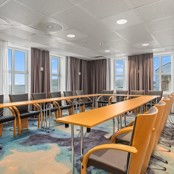 Konferanserom med U-formede trebord og polstrede stoler, arrangert for møter. Store vinduer slipper inn naturlig lys. Teppet er mønstret i blå og beige toner.
