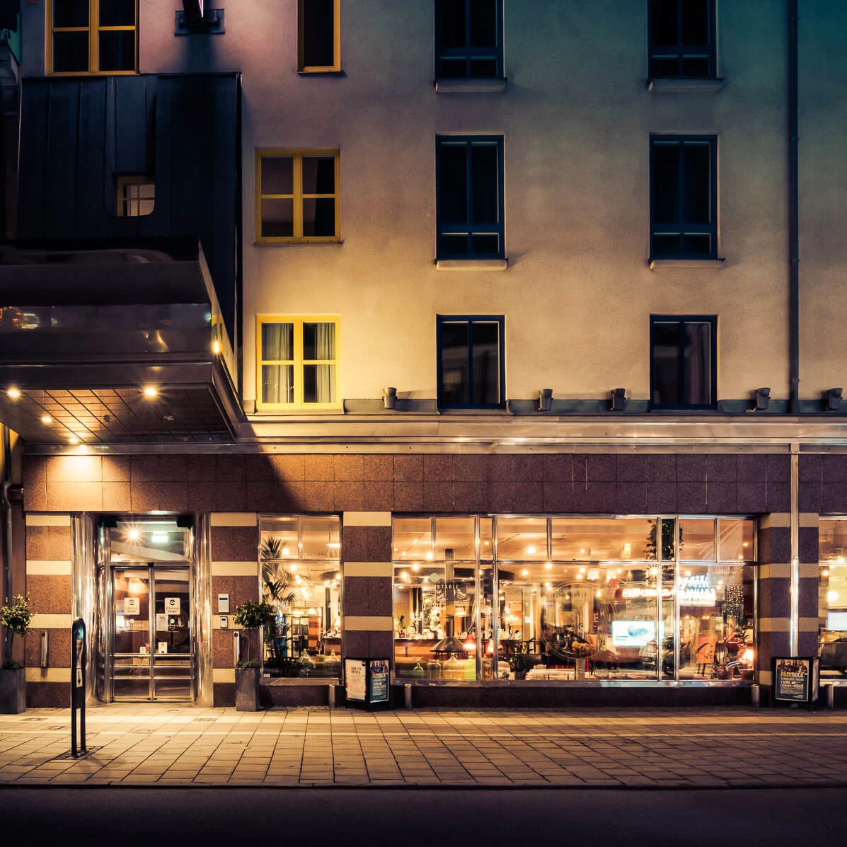 A building facade, illuminated at night, showcases large windows revealing a warmly lit interior with people inside. A sign with "Göpik" is visible to the left.