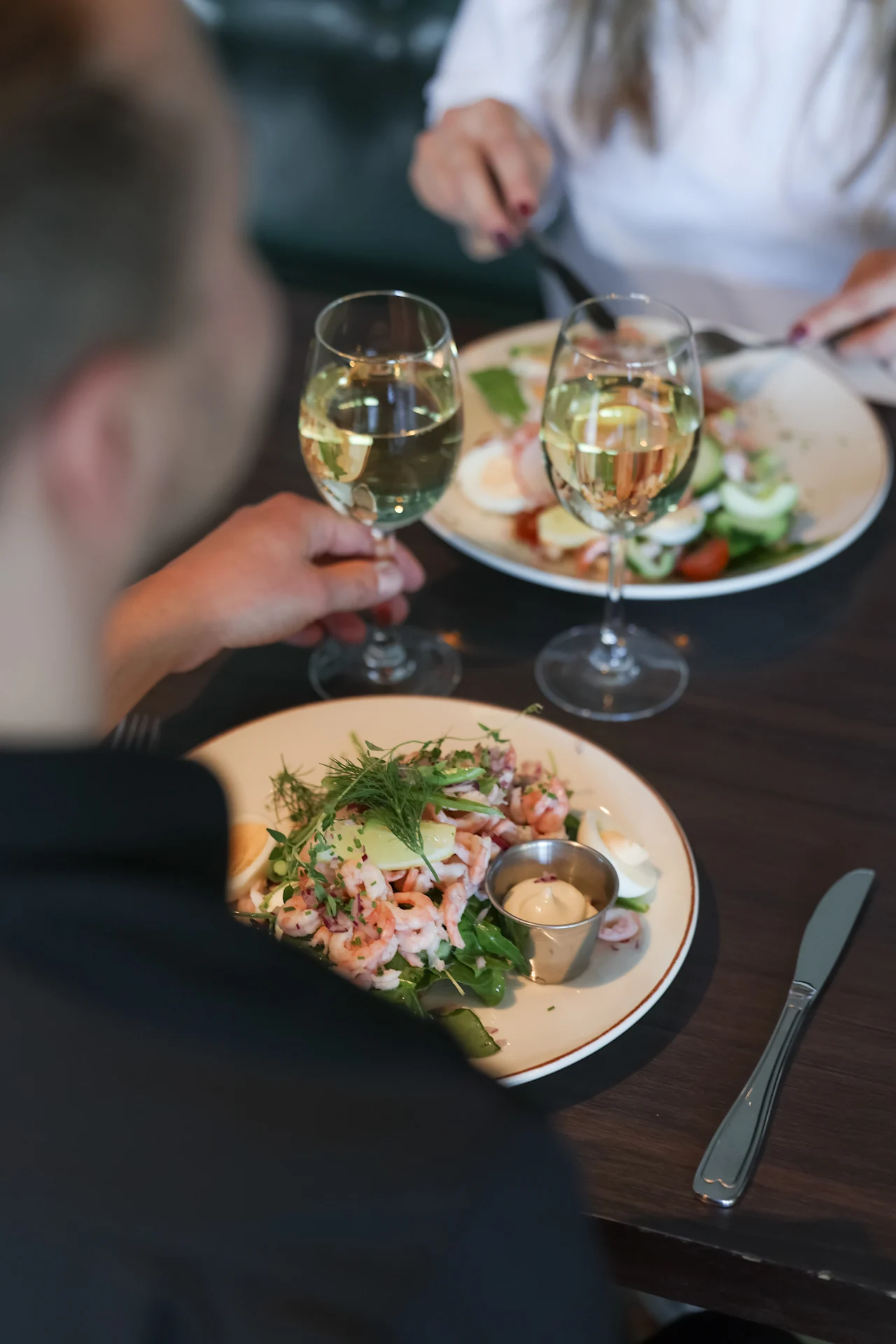 A seafood salad garnished with herbs is served on a white plate, accompanied by a glass of white wine, on a wooden dining table set for two people.