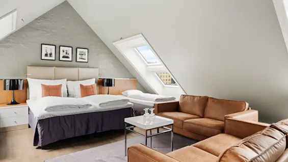 Et soveværelse med en stor seng og puder samt en brun sofa, under et skrånende loft med ovenlysvinduer. Indrammede billeder pryder væggen over sengen.
