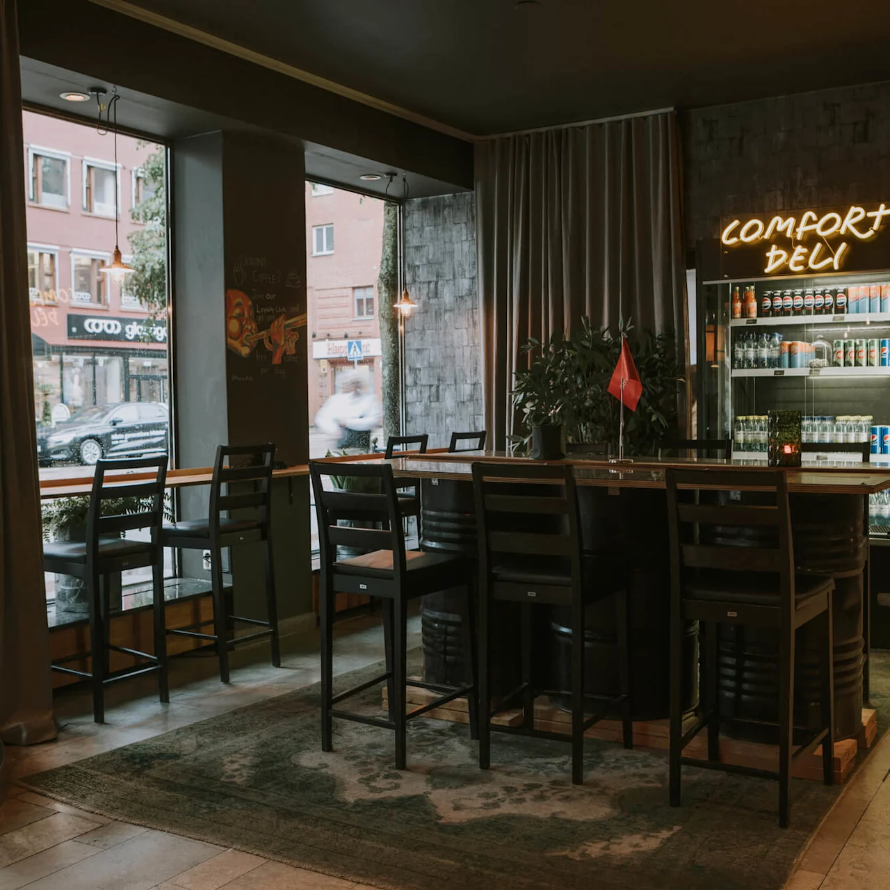 Tomme barborde langs et vindue, kigger ud over en bygade, neonskilt siger "COMFORT DELI" i et hyggeligt, dæmpet belyst interiør.