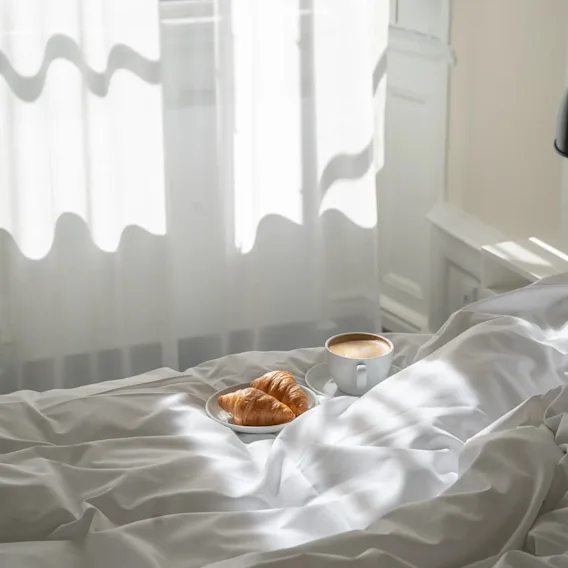 Morgenmad på sengen med croissanter og kaffe, sollys strømmer ind gennem hvide gardiner.