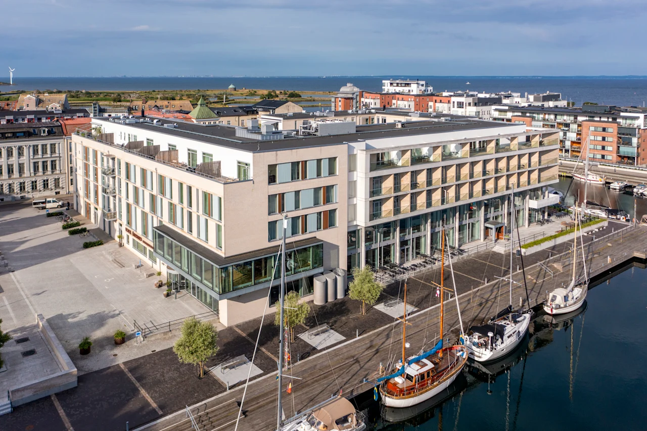 Moderni hotellirakennus lasijulkisivulla, josta on näkymä satamaan, jossa on useita veneitä, kaupunki ja meri taustalla.